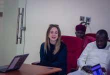 Three people seated, one woman smiling, two men beside her, one using a phone. A laptop is open on the table.