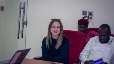 Three people seated, one woman smiling, two men beside her, one using a phone. A laptop is open on the table.
