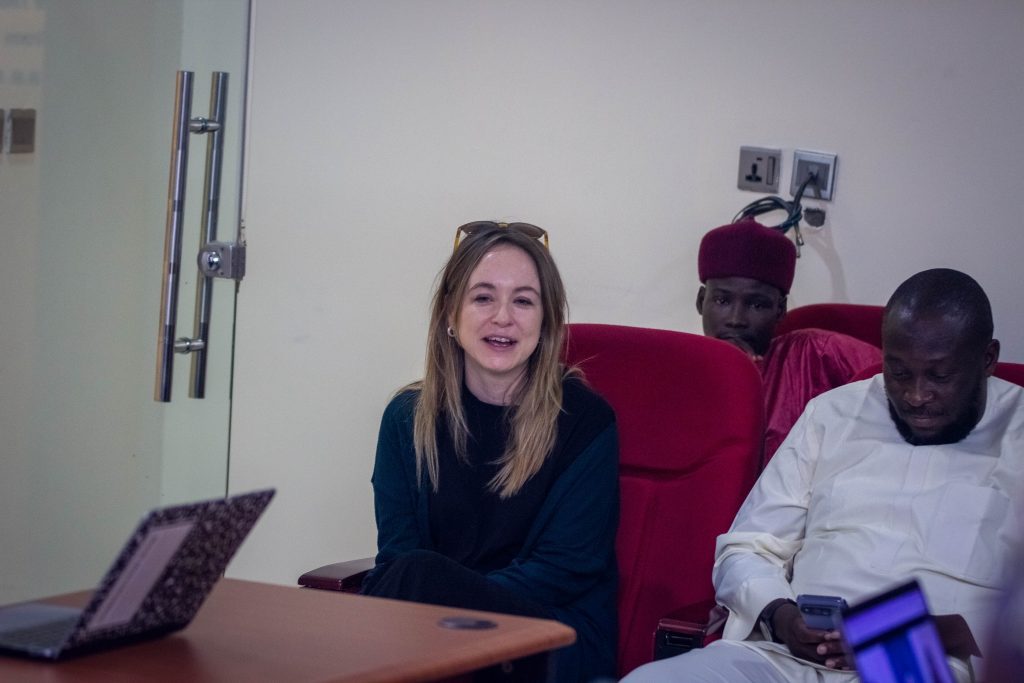 Three people sitting in a room; a woman smiling and two men beside her, with one using a smartphone, and a laptop in the foreground.