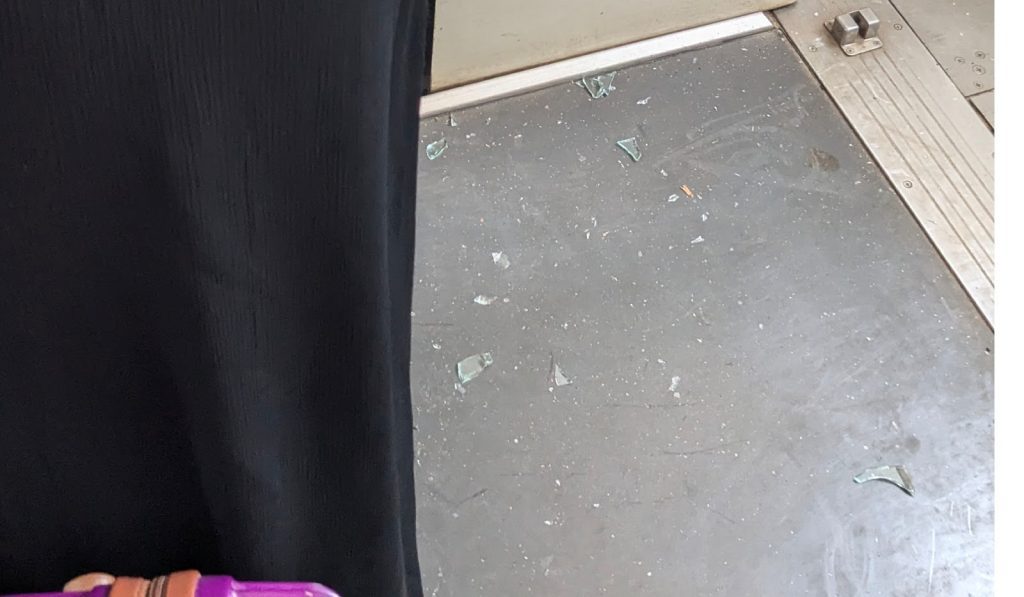 Broken glass pieces on a train floor near a black curtain and a person's hand holding a purple object.
