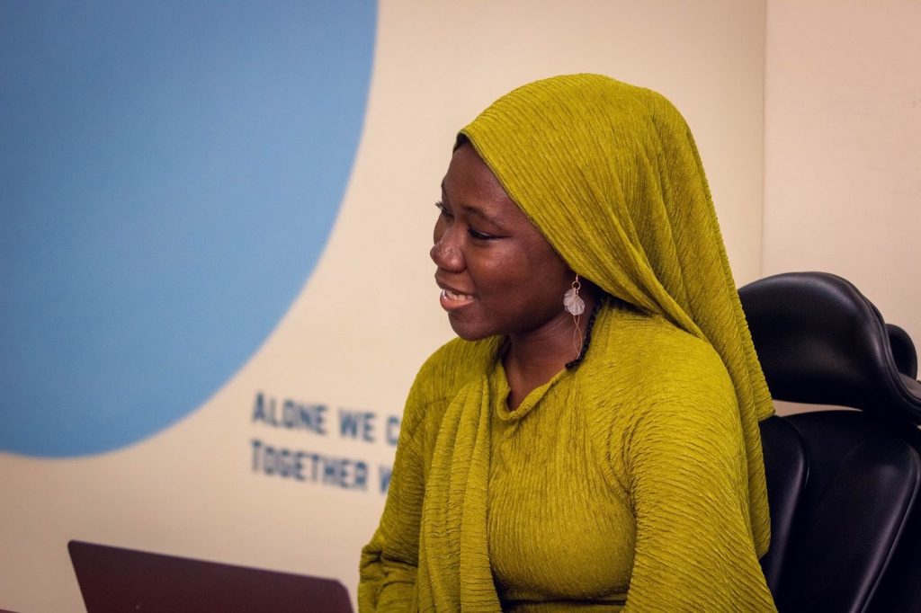 A woman in a green headscarf smiles while seated in front of a partially visible sign, with a blue circle in the background.
