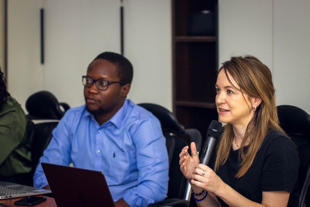 Two people in a meeting room: a woman speaking into a microphone and a man listening attentively, both with laptops in front of them.
