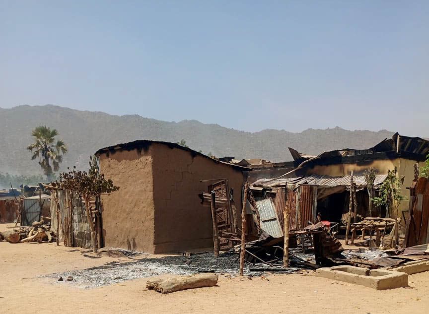 Burnt-out structures with metal roofs in a rural area, set against a backdrop of mountains and clear skies.