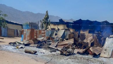 Collapsed corrugated metal structures with visible burn marks and smoke, set against a backdrop of mountains and clear blue sky.