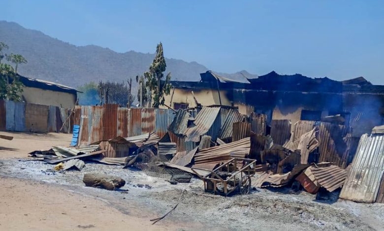 Collapsed corrugated metal structures with visible burn marks and smoke, set against a backdrop of mountains and clear blue sky.