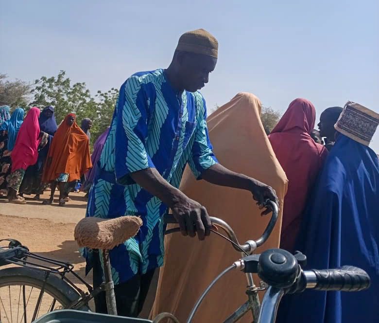 Man in blue shirt with bicycle stands near a line of people in colorful clothing under a clear sky.