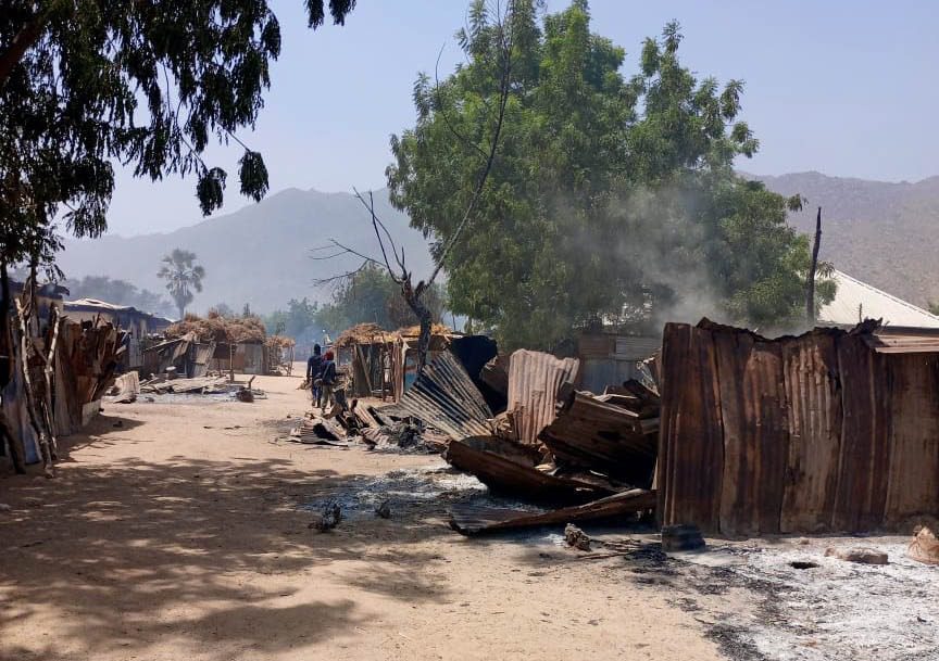 A dirt street lined with damaged, charred structures, and scattered debris, with smoke rising and a tree in the background.