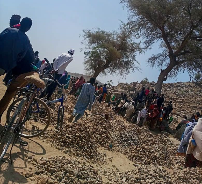 A group of people gather in a rocky, arid area with trees, some with bicycles, standing and walking around under a clear blue sky.