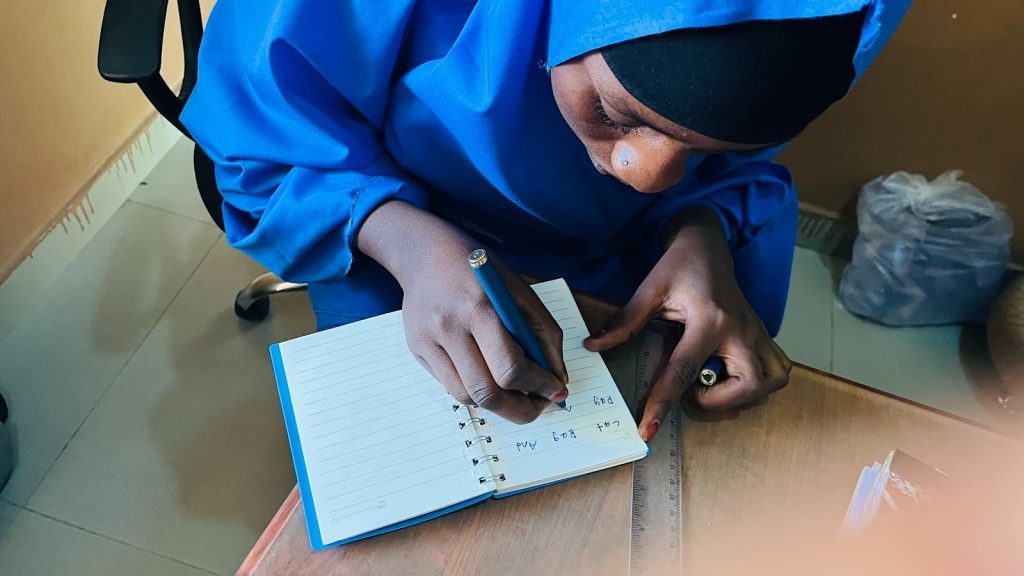 Person in blue clothing writing in a notebook on a table, with a ruler beside them.