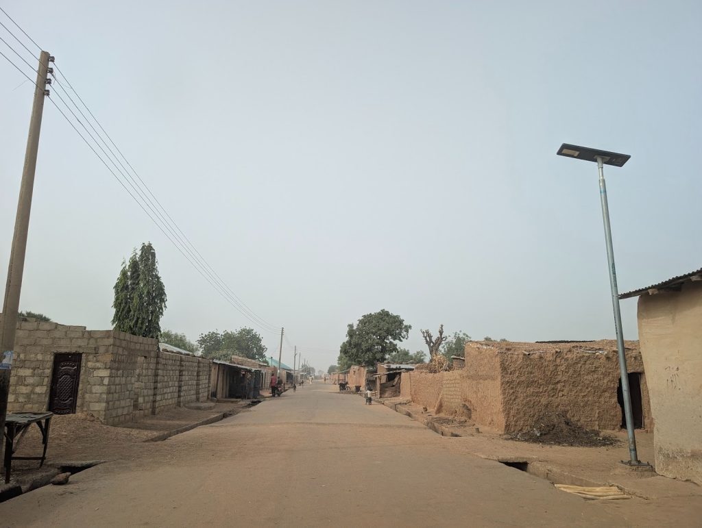 A dirt road flanked by rustic buildings and trees, with utility poles lining the street under a clear sky.