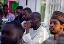 People seated at a gathering, with one person attentively listening, hands clasped near their face.