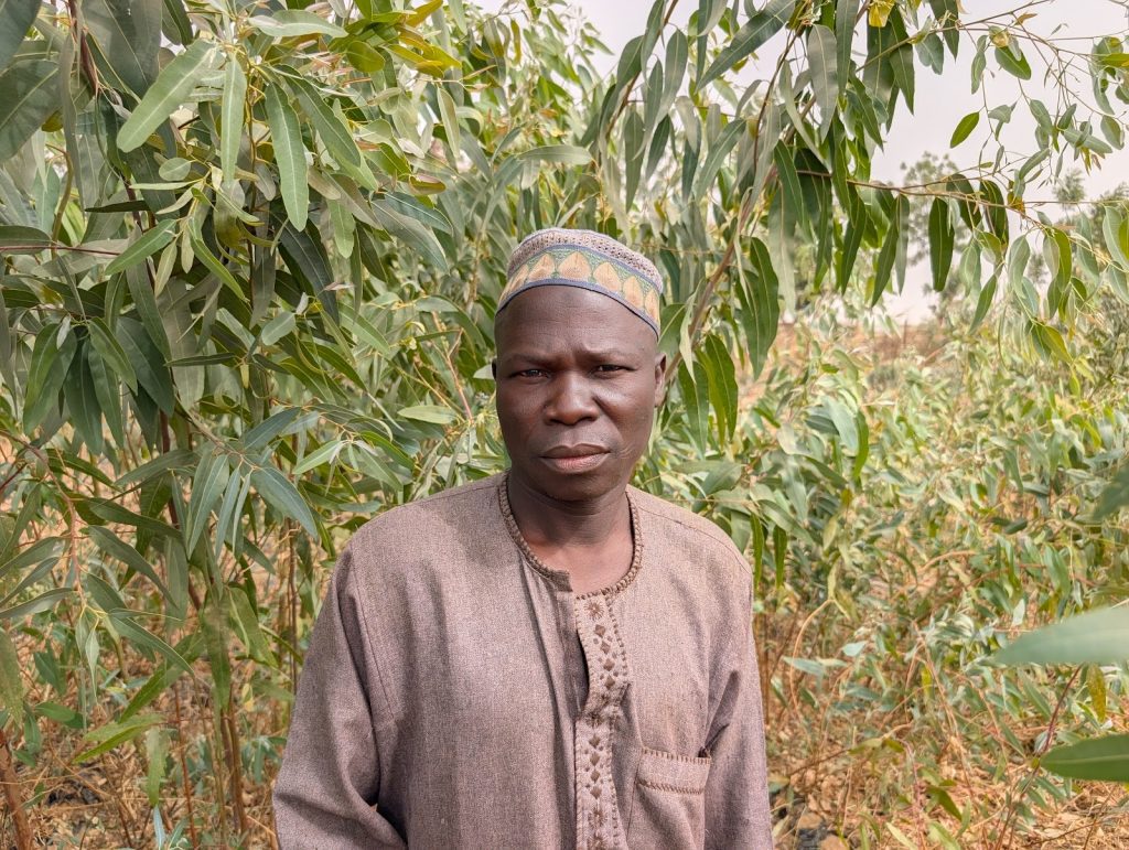 Man in traditional attire stands amidst lush green foliage.