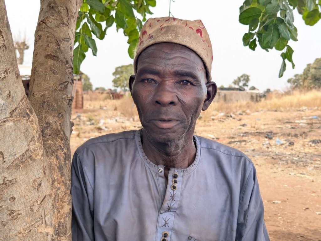Elderly man in traditional attire and hat stands beside a tree, with a rural background in view.