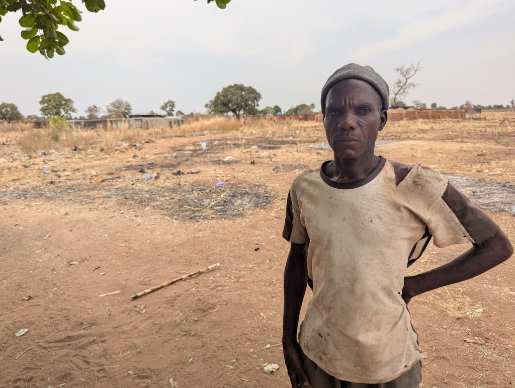 A man in a worn-out shirt stands in a dry, open field with scattered trees and debris under a cloudy sky.