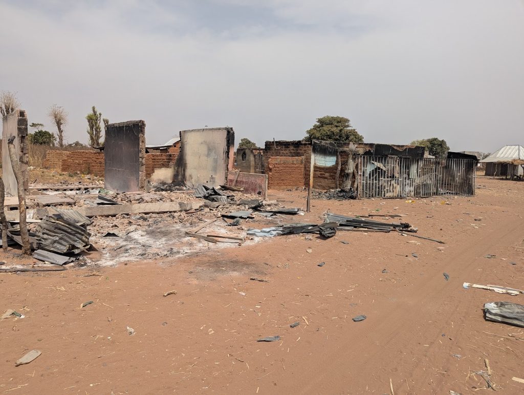 A heavily damaged building with charred walls and scattered debris in a barren landscape under a hazy sky.