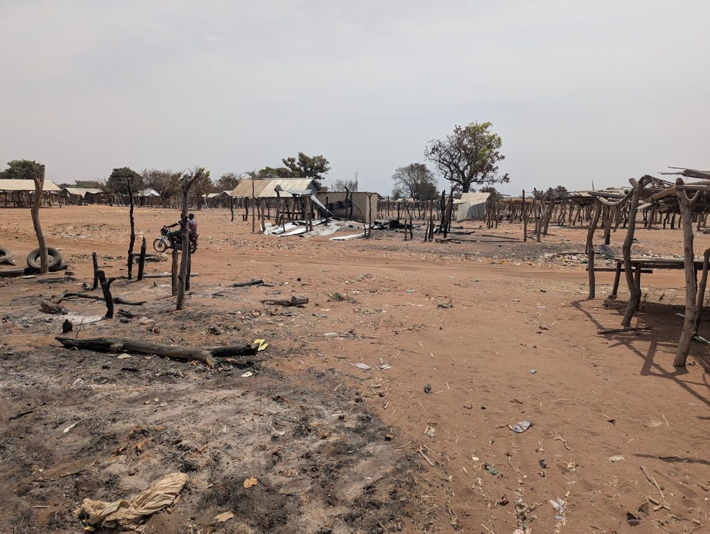 Deserted, charred structures and debris in a dusty landscape, with a person on a motorcycle in the background under a clear sky.