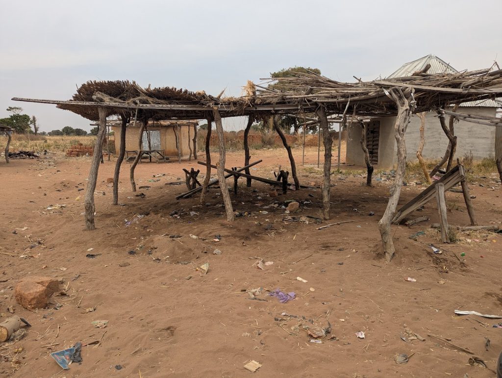 Dilapidated wooden shelter with thatched roof on a dusty field, surrounded by scattered debris and a few distant trees.
