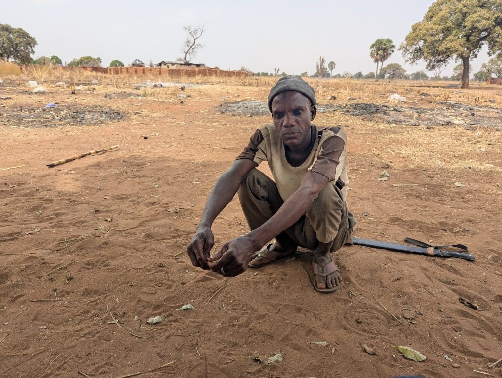 Man squatting on dry ground in a rural area, with trees and a building in the background.