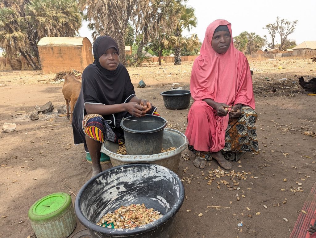 Two women sitting on the ground, shelling nuts into bowls, with dry landscape and simple huts in the background.