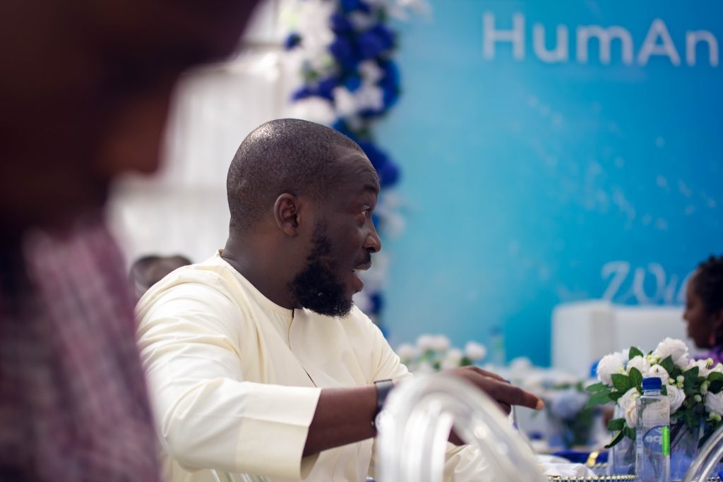 A man in a white shirt speaks animatedly at an event, with blue and white decor in the background.