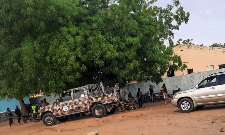 Camouflage vehicle and silver SUV parked under a large tree with people and motorcycles nearby.