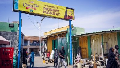 People walk under a "Welcome to Monday Market" sign in an outdoor market with colorful buildings and stalls.