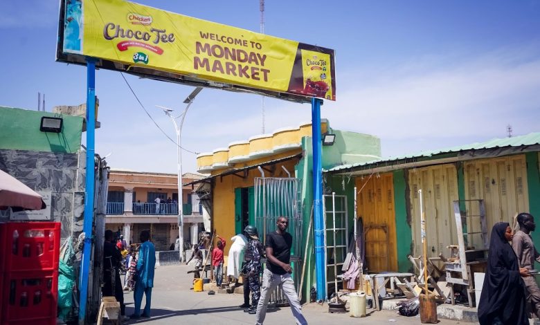 People walk under a "Welcome to Monday Market" sign in an outdoor market with colorful buildings and stalls.