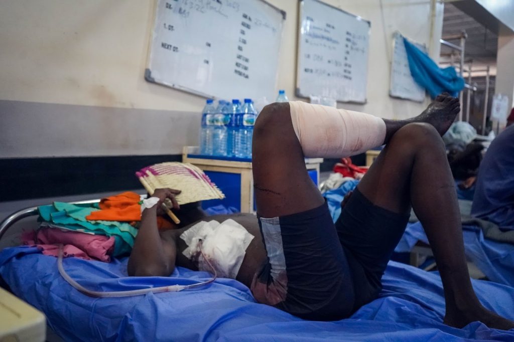 Person lying on hospital bed with leg bandaged, holding a fan; medical tubes visible. Boards with writing in the background.