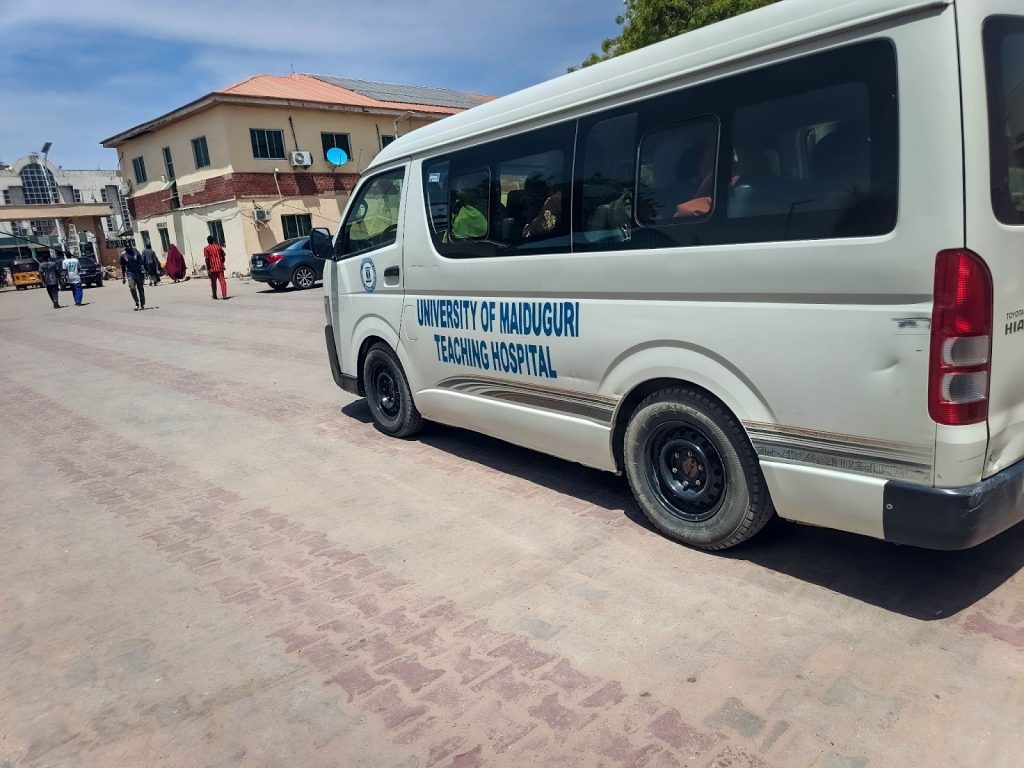White van with "University of Maiduguri Teaching Hospital" parked outside a building under a clear sky. People walking in the background.