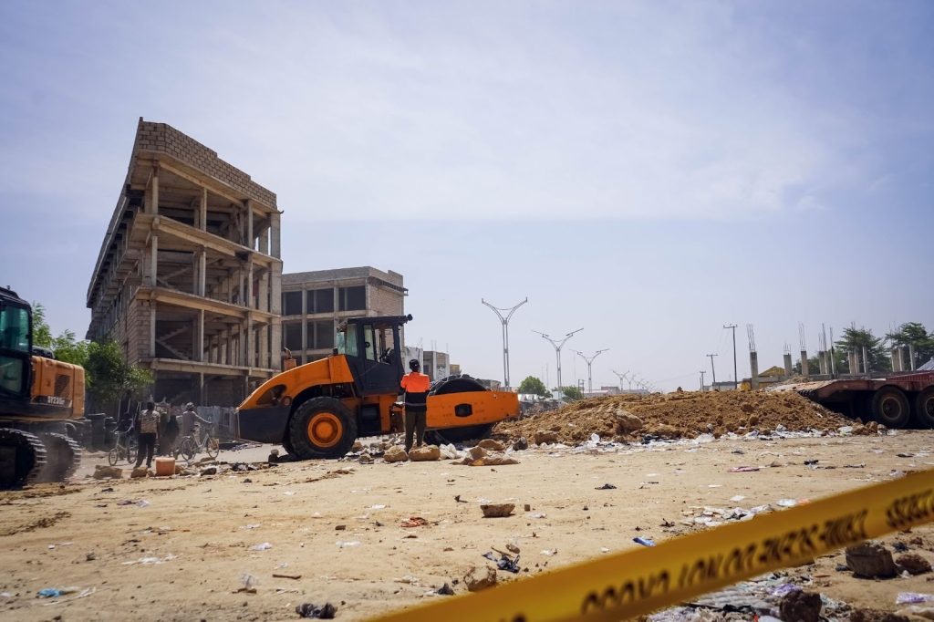 Construction site with heavy machinery, debris, and an unfinished building. Worker in orange vest near a steamroller. Caution tape in foreground.