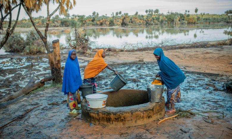 Three people gather water from a well beside a river, surrounded by trees and muddy ground.