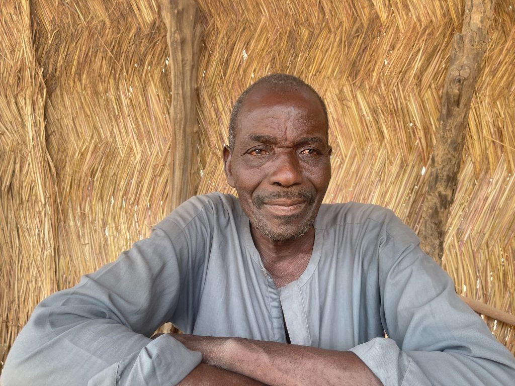 Man in a gray shirt sits relaxed against a thatched background, looking at the camera with a calm expression.