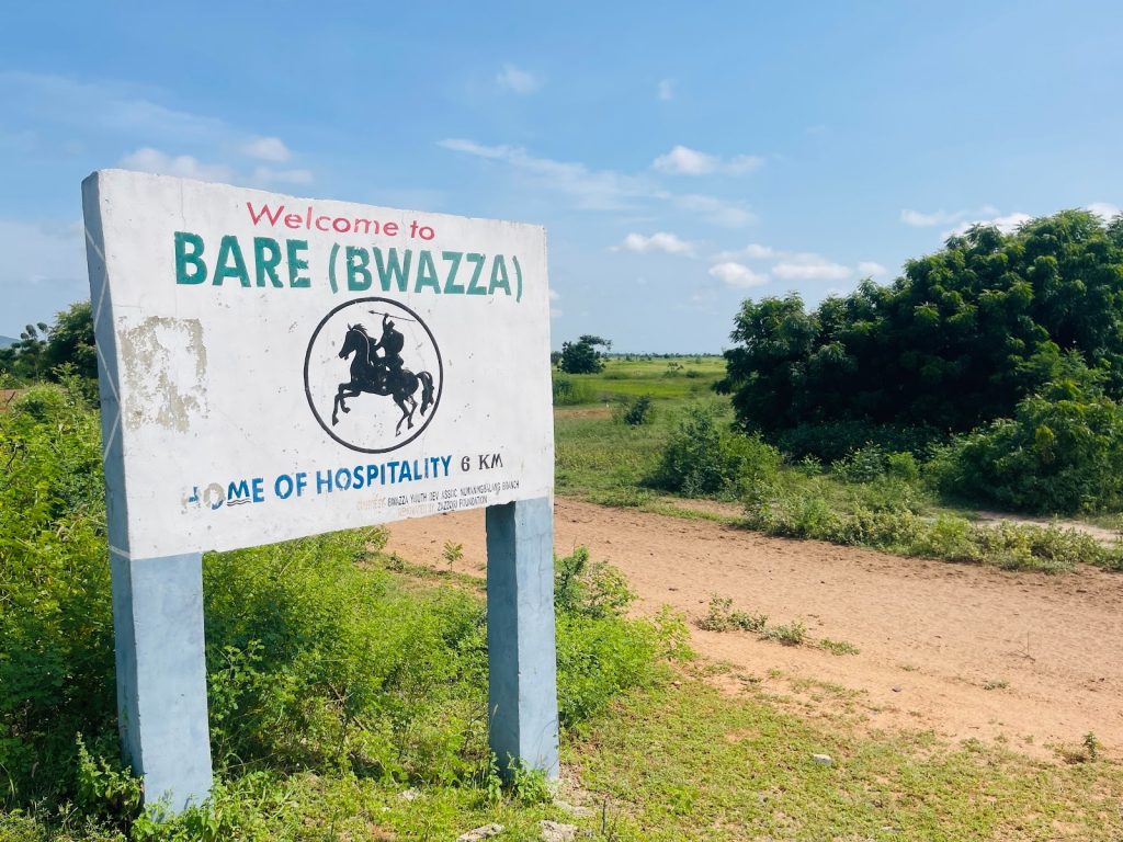 Sign reading "Welcome to Bare (Bwazza), Home of Hospitality" near a dirt road and greenery under a clear blue sky.