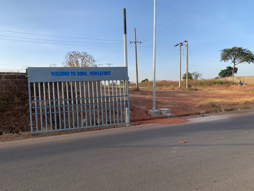 Gate with "Welcome to Dong, New Layout" sign beside a grassy field and utility poles under a clear sky.