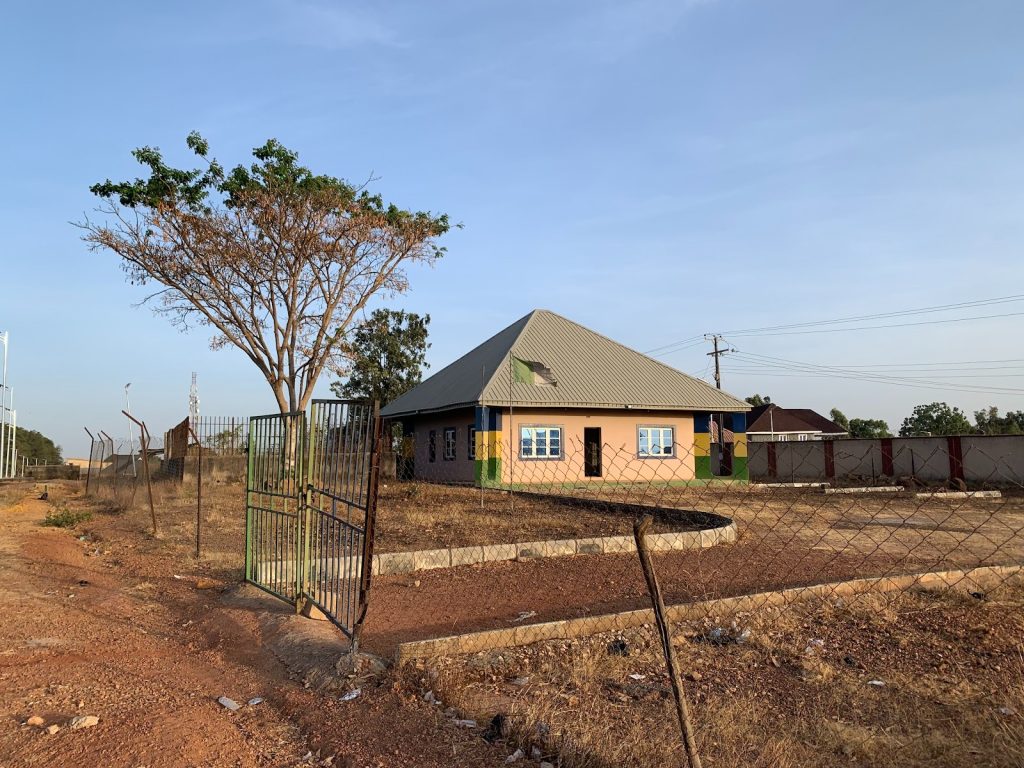 A house with a gray roof and blue windows stands behind a wire fence and open gate, with a tree on a dirt path under a clear sky.