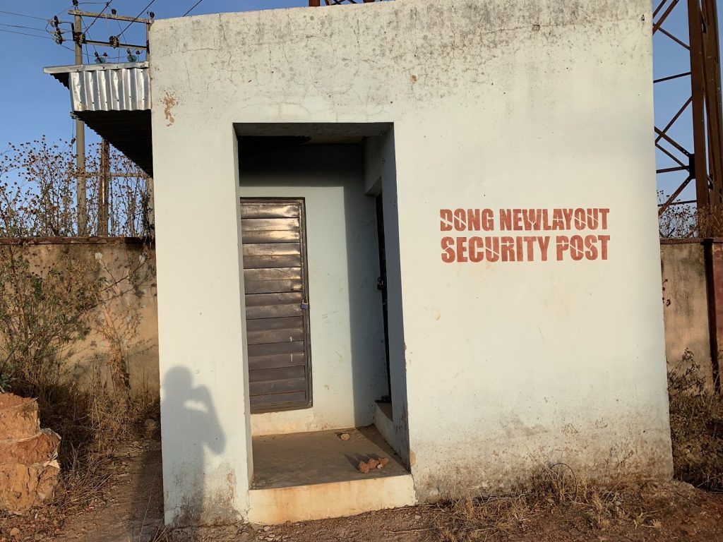 Small white security post with a door, near a fence. Text on the wall reads: "Dong New Layout Security Post."