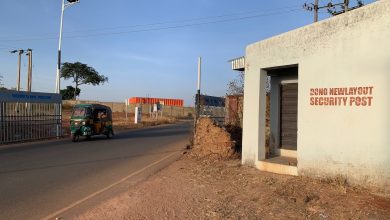 A tuk-tuk drives past a security post labeled "DONG NEW LAYOUT SECURITY POST" on a sunny day, with a gate in the background.