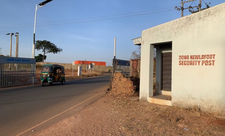 A tuk-tuk drives past a security post labeled "DONG NEW LAYOUT SECURITY POST" on a sunny day, with a gate in the background.