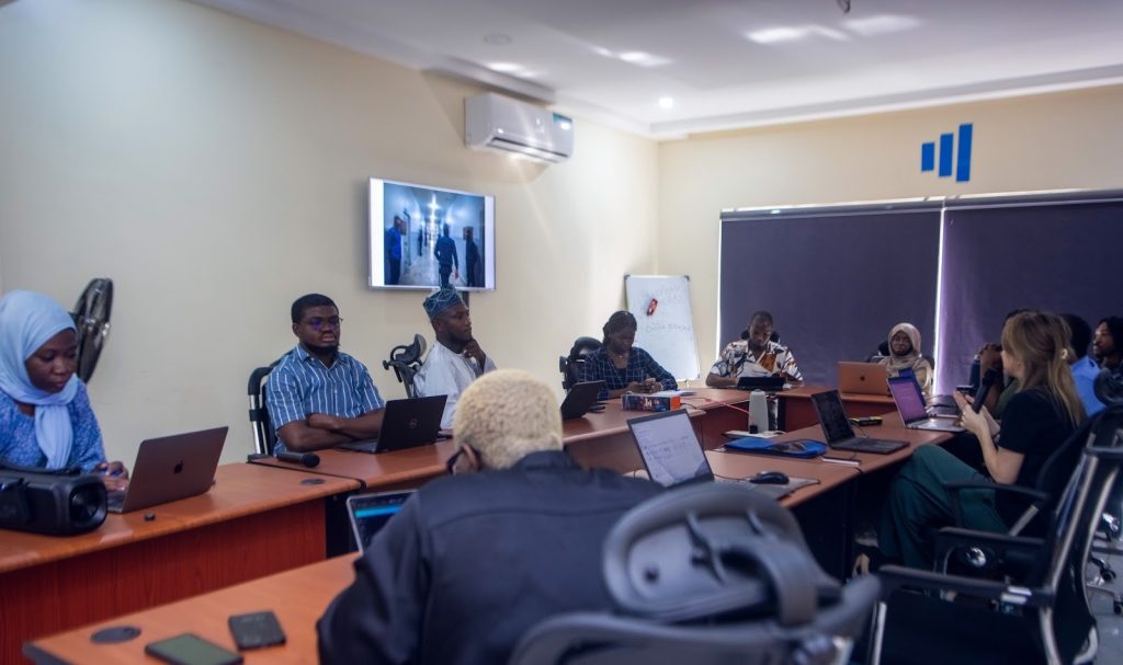 People sit around a conference table with laptops and papers during a meeting in an office setting.