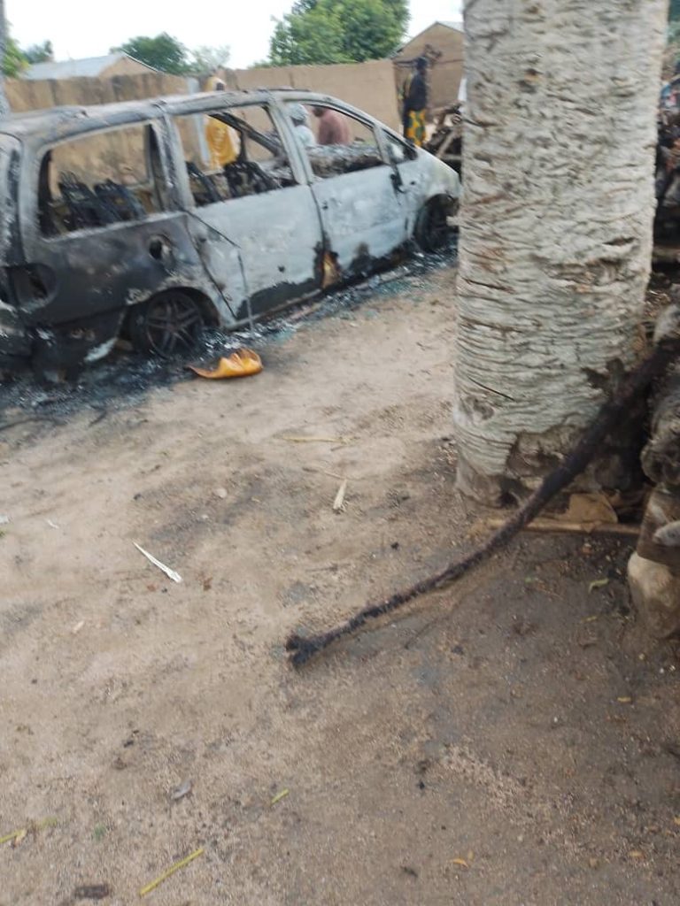 Burnt-out car on a dirt road with two people nearby and a tree in the foreground.