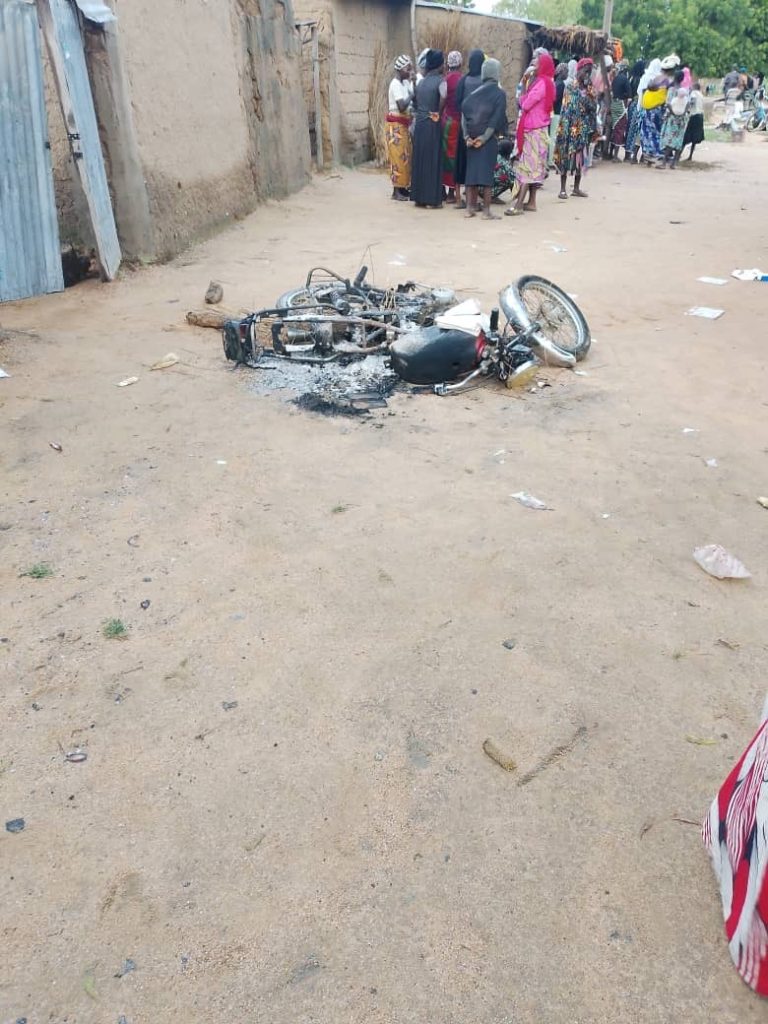 A burned motorcycle lies on the ground in a dirt alley. A group of people stand in the background, gathered in a discussion.