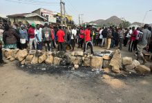 A large crowd gathers near a barricade of rocks on a street, with hills visible in the background. Some buildings and power lines are nearby.