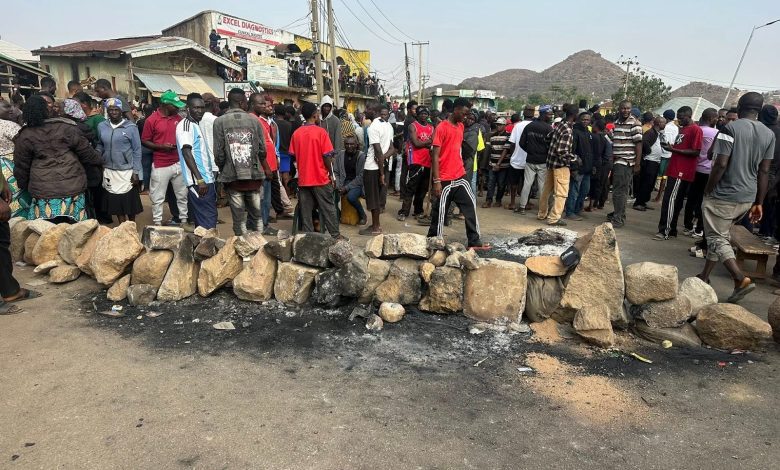 A large crowd gathers near a barricade of rocks on a street, with hills visible in the background. Some buildings and power lines are nearby.