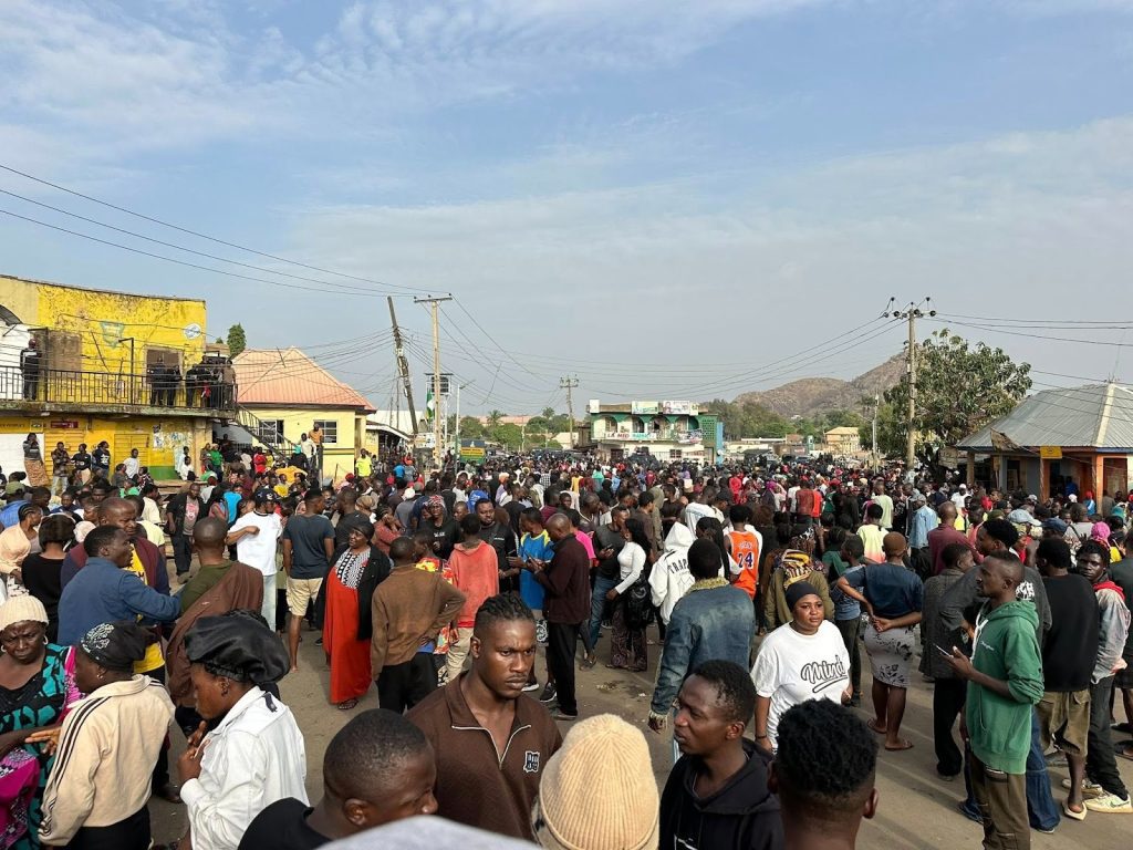 Crowd gathered in a street surrounded by buildings, trees, and hills in the background under a clear sky.