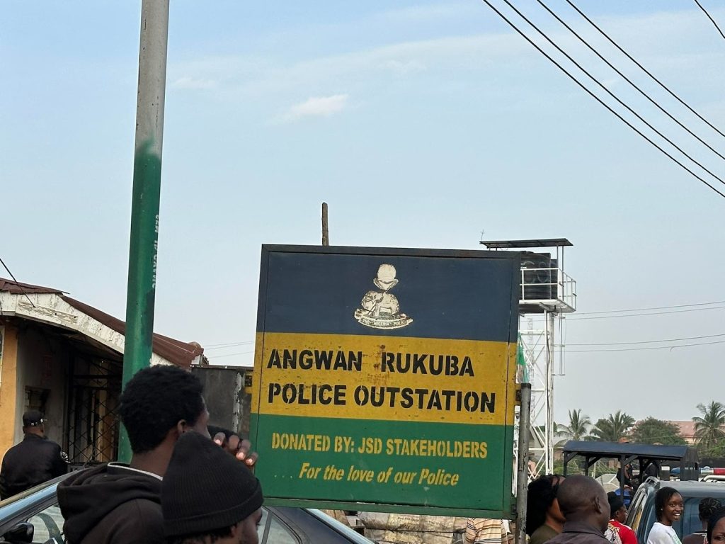 Sign for Angwan Rukuba Police Outstation with people nearby. Text mentions JSD Stakeholders' donation for love of police.