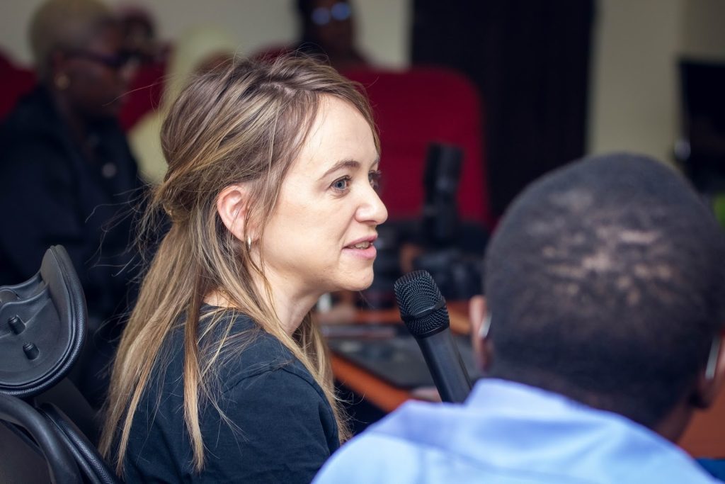 A woman speaking into a microphone at an indoor event, with people in the background.