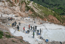 People working in a busy hillside mining area with scattered pools and equipment, surrounded by green landscape below.