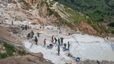 People working in a busy hillside mining area with scattered pools and equipment, surrounded by green landscape below.