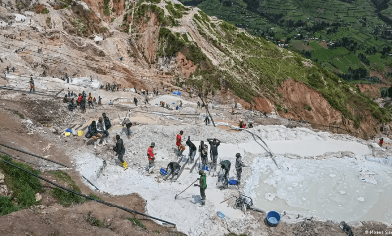 People working in a busy hillside mining area with scattered pools and equipment, surrounded by green landscape below.