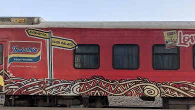 Red train car with Indomie noodle ad, colorful patterns, and street signs pointing to B.Gwari Rd. and Emir Palace.
